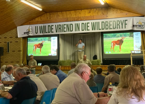 A group of people seated at tables in a hall with a wooden ceiling, watching a presentation. Two screens display an image of an animal, and a person is speaking on stage. A banner above reads "U Wilde Vriend in die Wildbedryf."